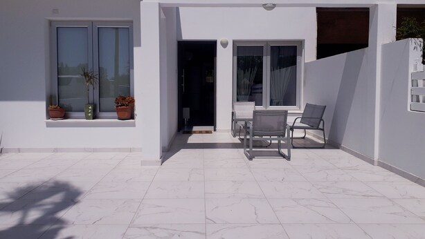 A modern building with a white exterior, a tiled patio, and a small seating area with two chairs. - Eva Christ Apt (Oroklini)