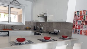 A modern kitchen with white cabinets, a central island with bar stools, a built-in oven, and a coffee machine. - Eva Christ Apt (Oroklini)