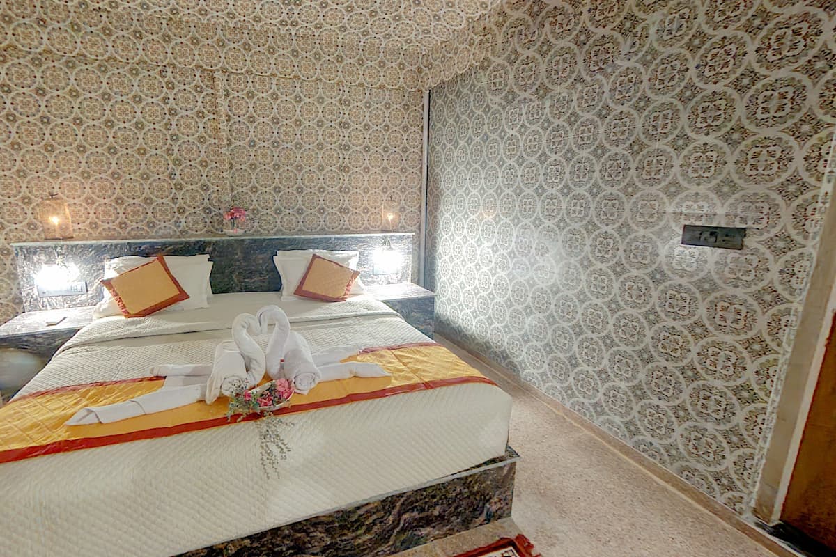Une chambre d’hôtel avec un papier peint à motifs, un lit recouvert d’une couvre-lit jaune et blanche, deux oreillers et une table de chevet avec une lampe.