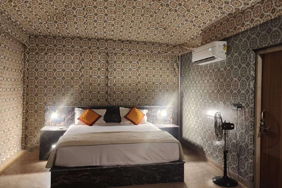 Une chambre avec un papier peint à motifs, un lit avec une literie blanche et des coussins orange, un ventilateur sur pied et un appareil de climatisation.
