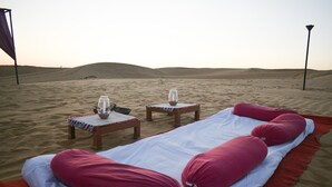A desert setting with a bed, pillows, a table with a vase, and a lamp post.