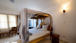 A four-poster canopy bed with white linens, a wooden nightstand, a bench, and a wall-mounted light fixture. - Nyali Villa (Mombasa)