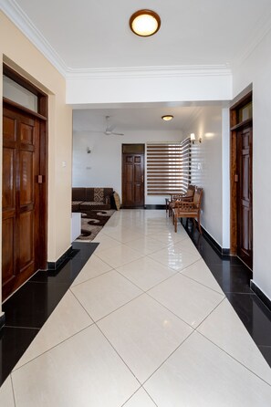 A hallway with a tiled floor, wooden doors, a ceiling fan, and a seating area with a patterned sofa and chairs.
