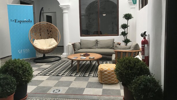 Un rincón interior acogedor con una silla de mimbre, un sofá y una mesita de centro. Hay un ventanal grande, una pared azul con un cartel y plantas en macetas.