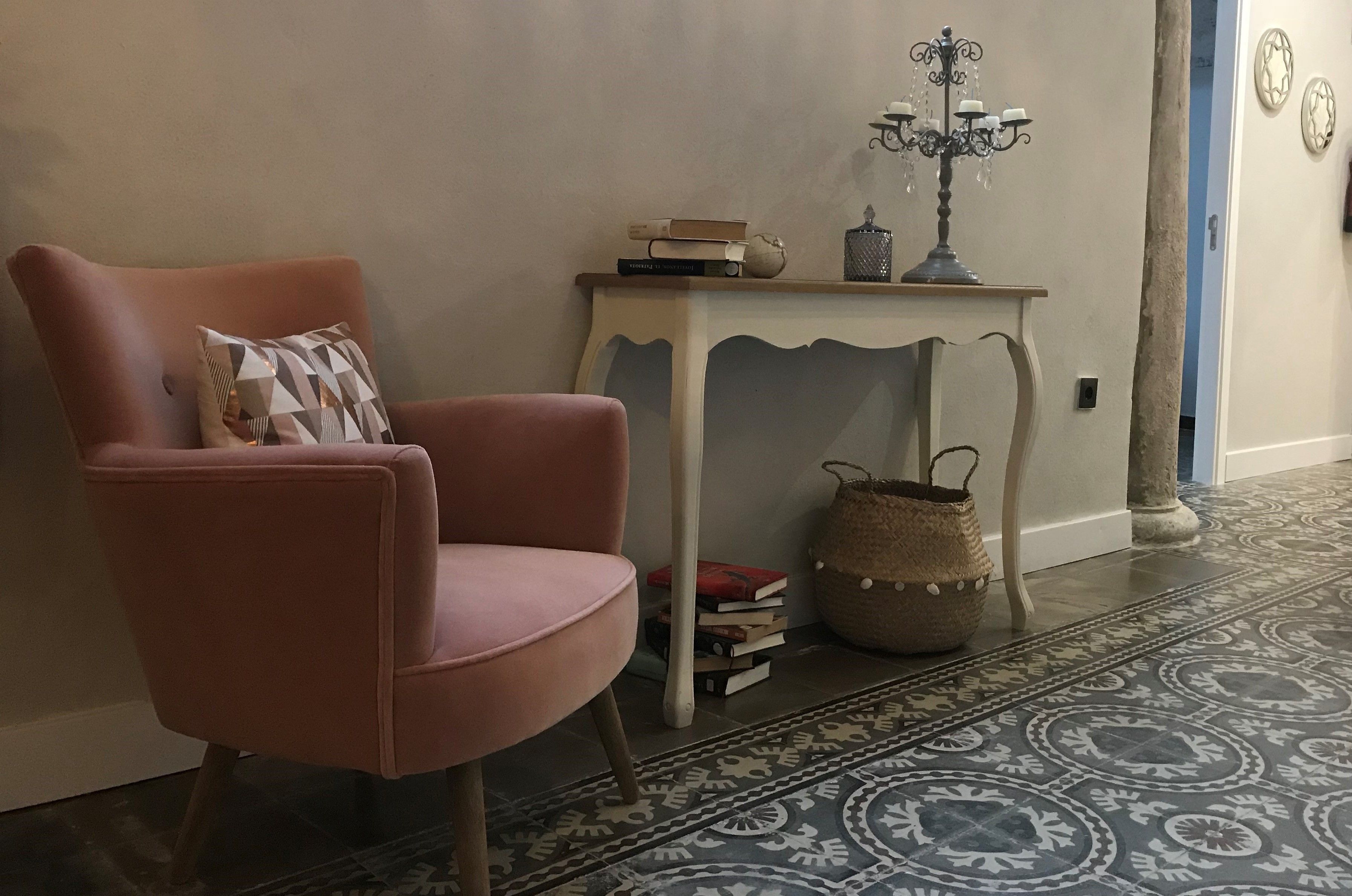 A pink armchair with a patterned pillow, a small white table with books and a candelabra, and a patterned rug.