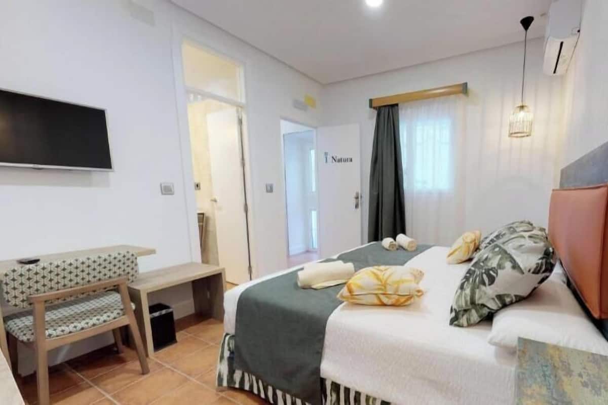 A hotel room with a bed, a desk, a television, and a door labeled 'Natura.'