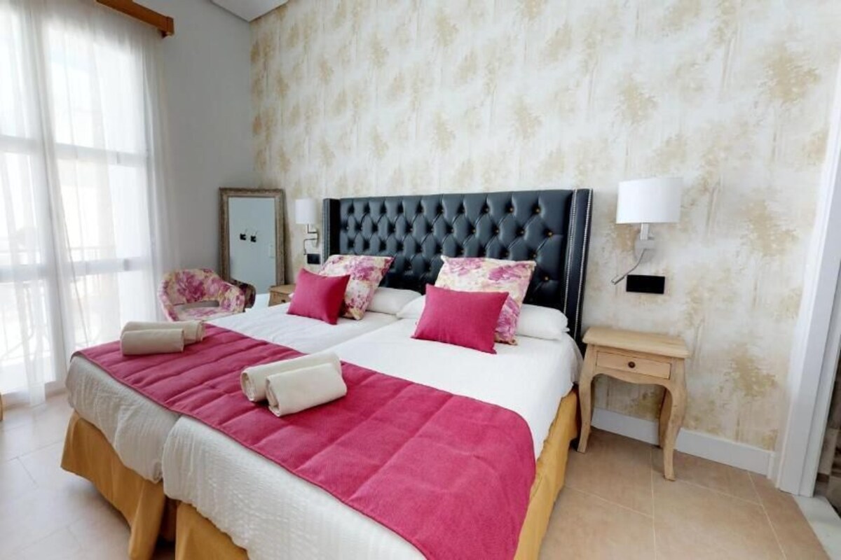 A double bed with a tufted headboard, two bedside tables, a mirror, and a window with curtains.