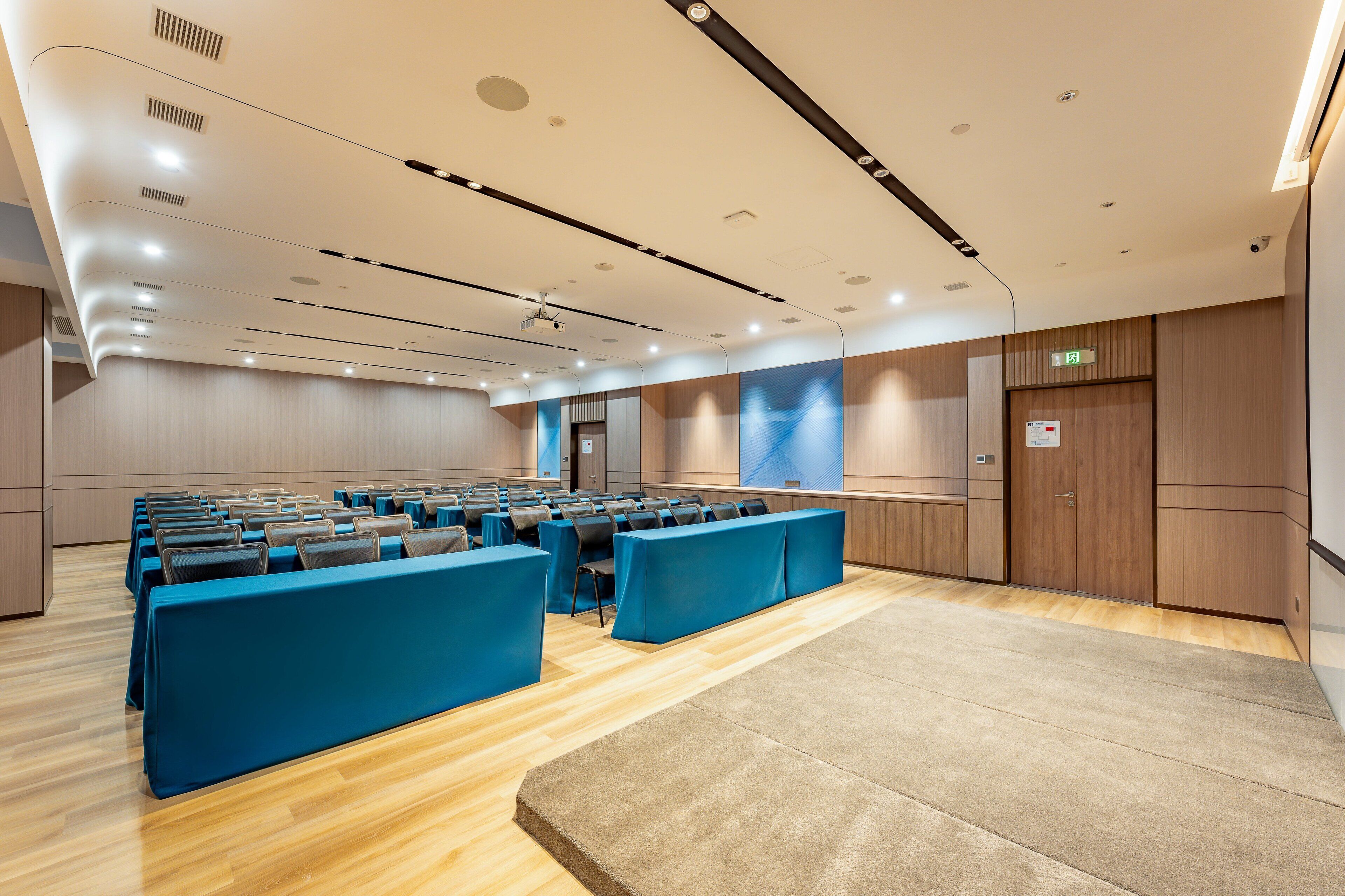 Un salón de conferencias con filas de mesas y sillas, suelo de madera, techo con iluminación empotrada y un proyector.