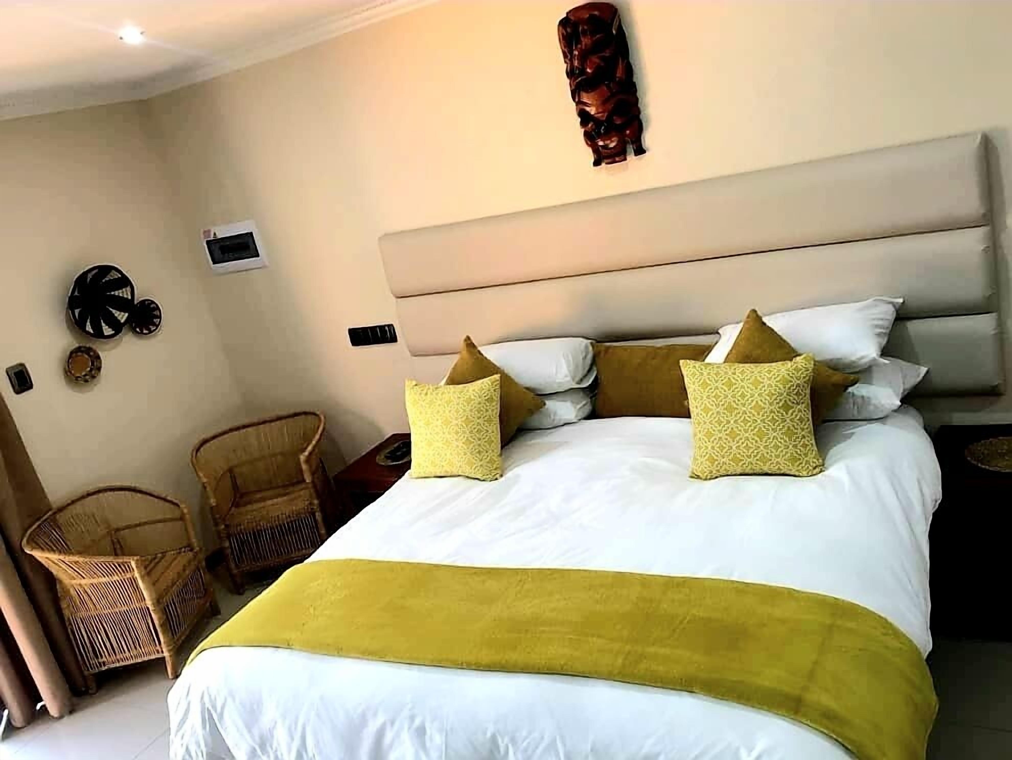 A hotel room with a large bed, a headboard, a wall-mounted artwork, a wicker chair, and a small table.