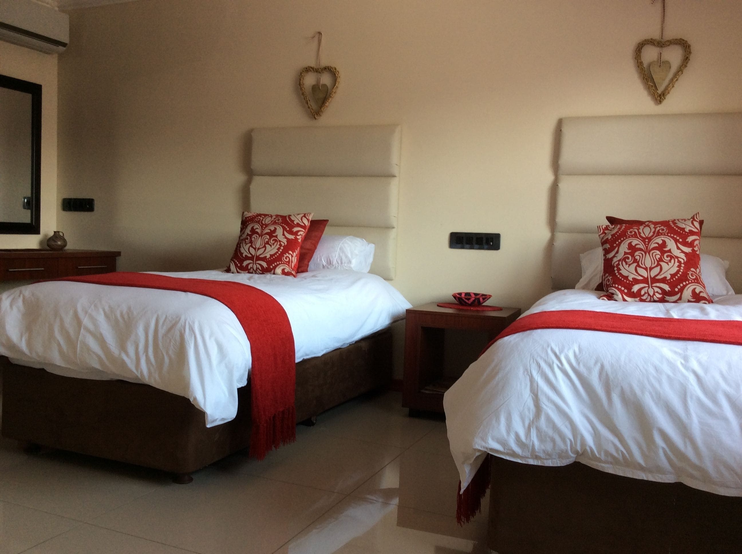 Two twin beds with white bedding and red accents, a small bedside table with a bowl, and heart-shaped wall decorations.