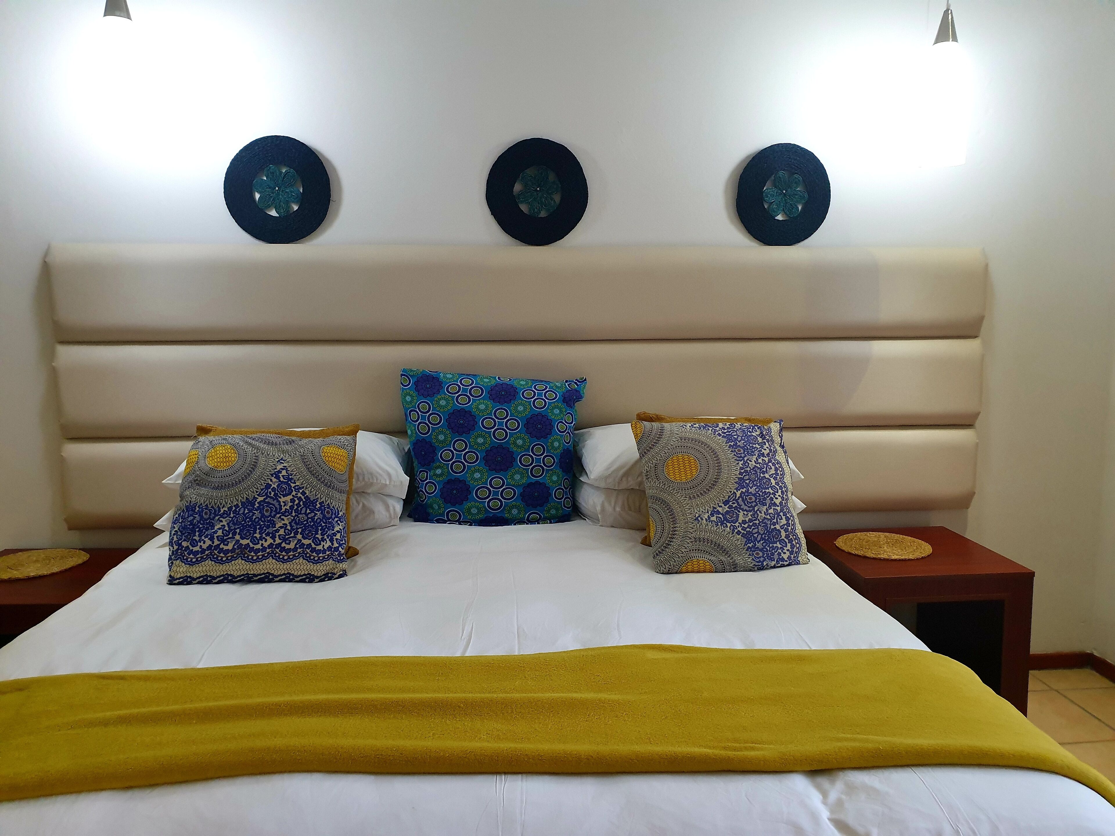 Un lit avec un drap blanc, un plaid jaune et trois coussins à motifs. Il y a deux tables de chevet avec des plateaux ronds, chacune surmontée d’une assiette décorative.