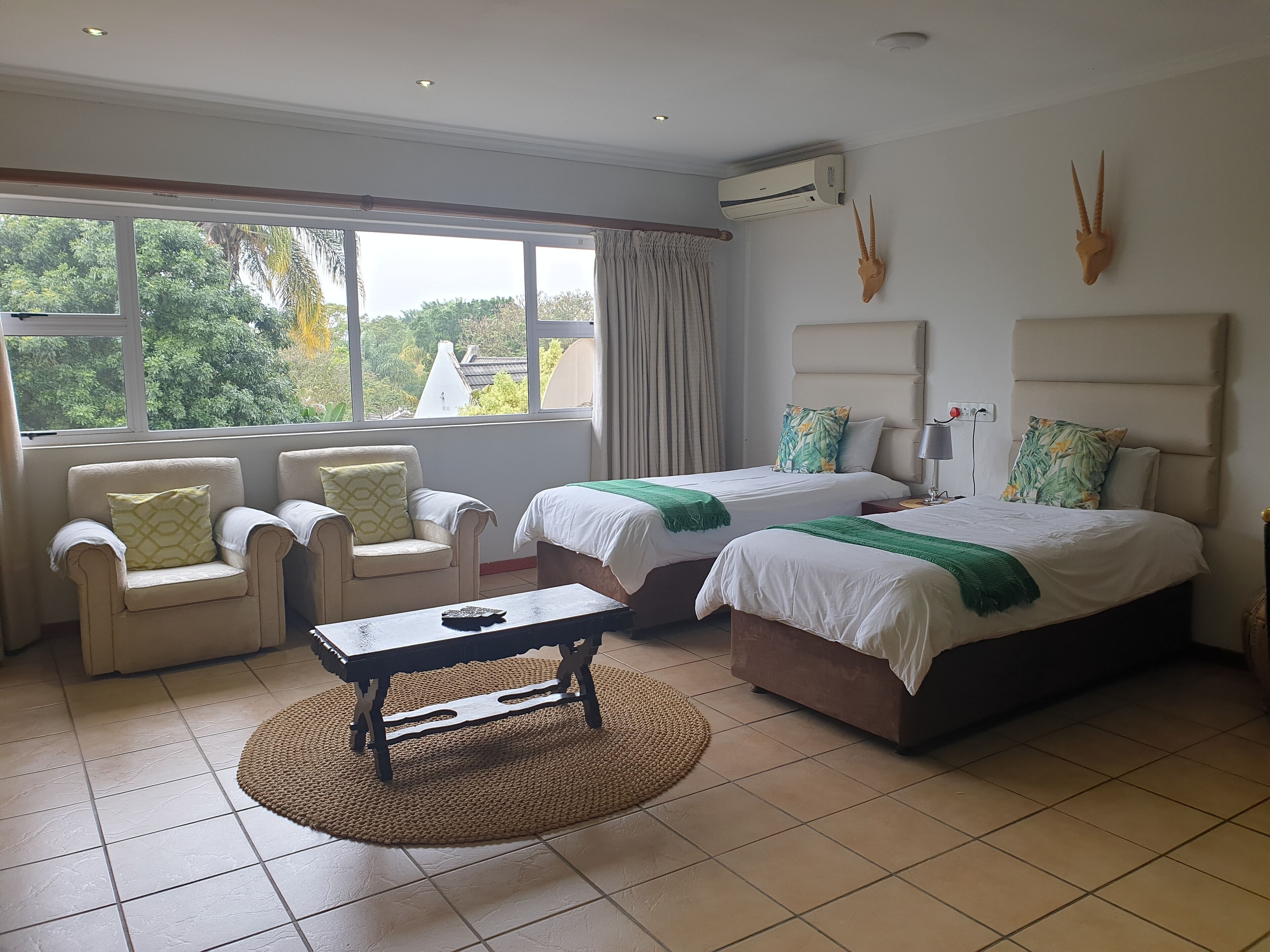 A hotel room with two beds, a sofa, a coffee table, and a view of greenery outside.