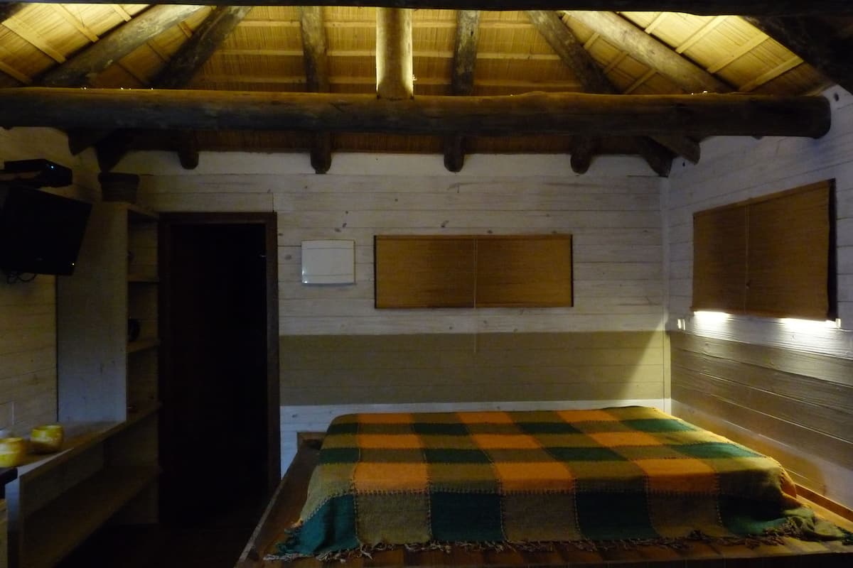 Una habitación con techo de madera, una cama con manta a cuadros, una repisa con velas y un interruptor de luz en la pared.