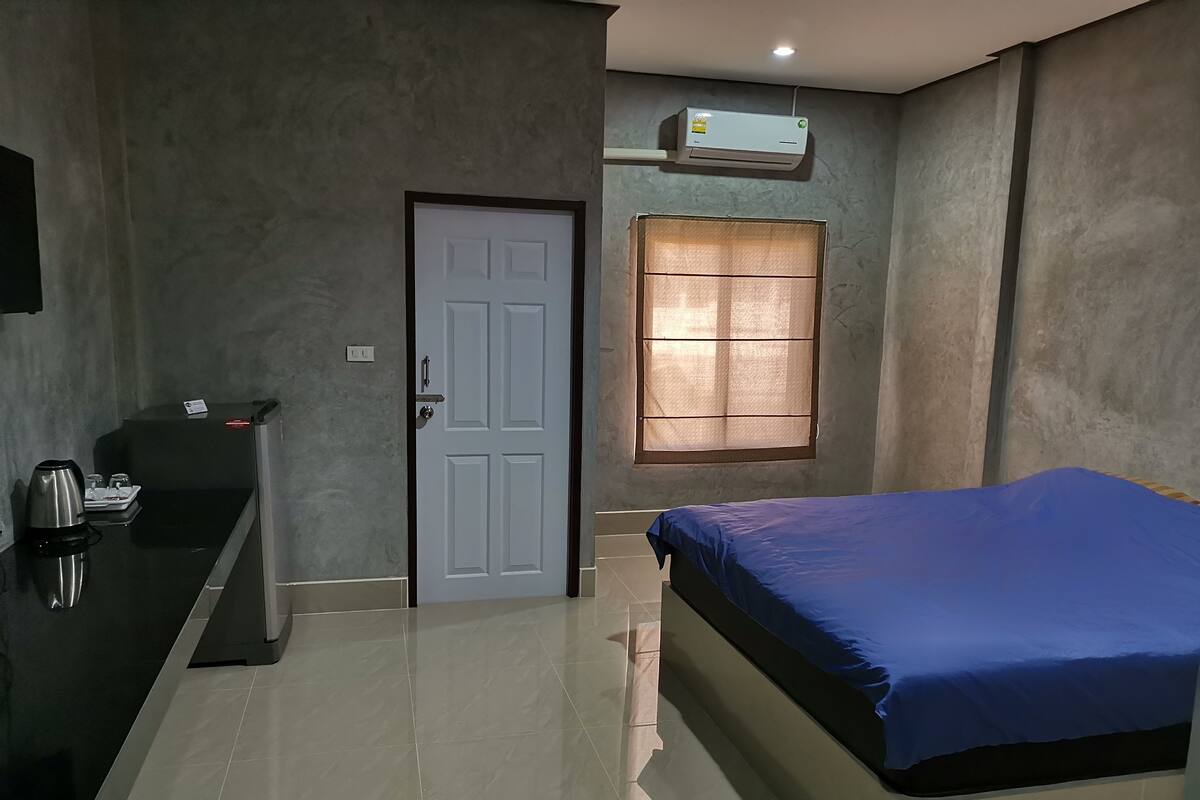 A room with a bed, a desk with a kettle, a door, and an air conditioning unit.