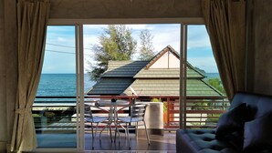A room with a large sliding glass door leading to a balcony with a view of the sea and a building. There is a table with chairs and a black sofa. - Sea Smile Resort (Prachuap Khiri Khan)
