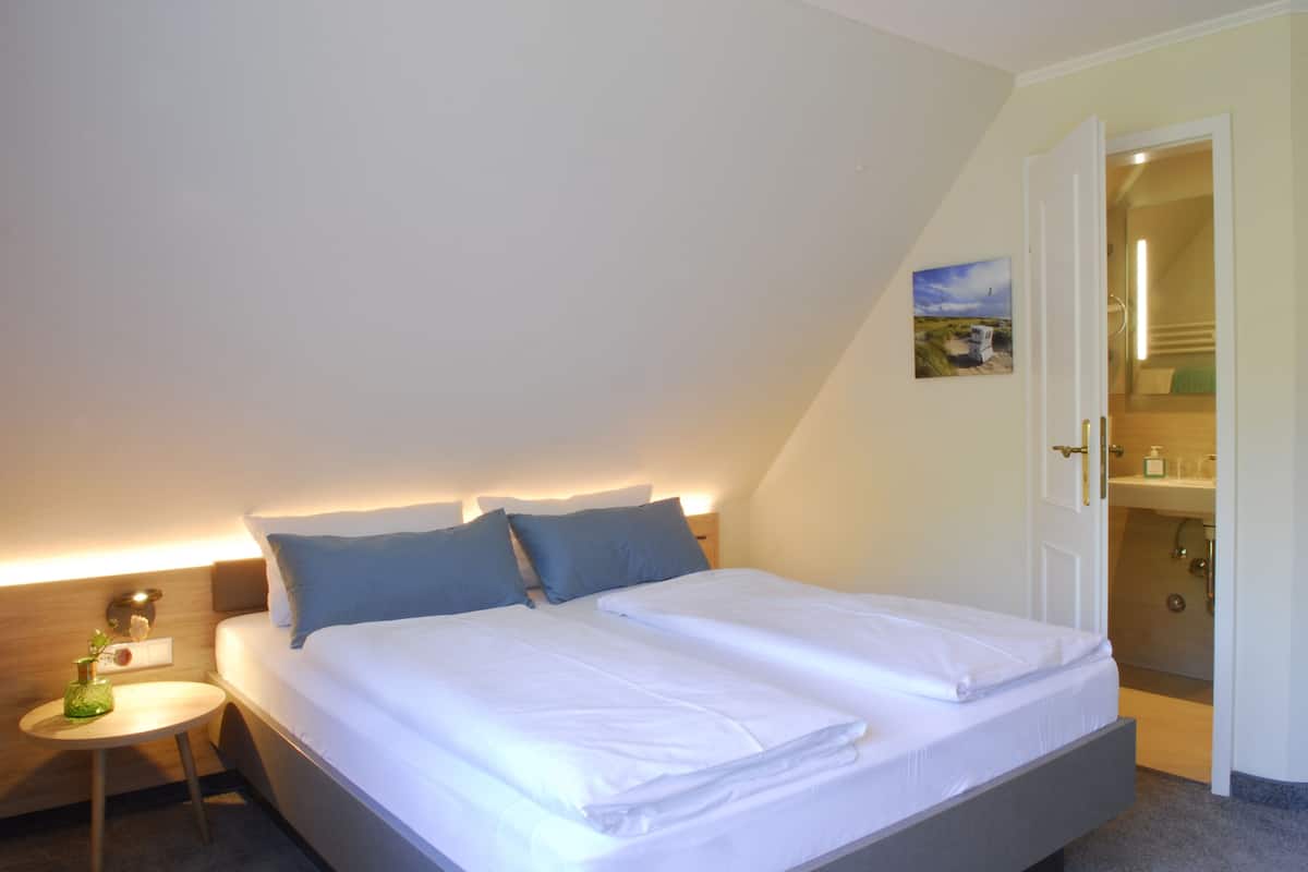 A neatly made twin bed with white linens and two pillows, a small round table with a vase, and a painting on the wall.