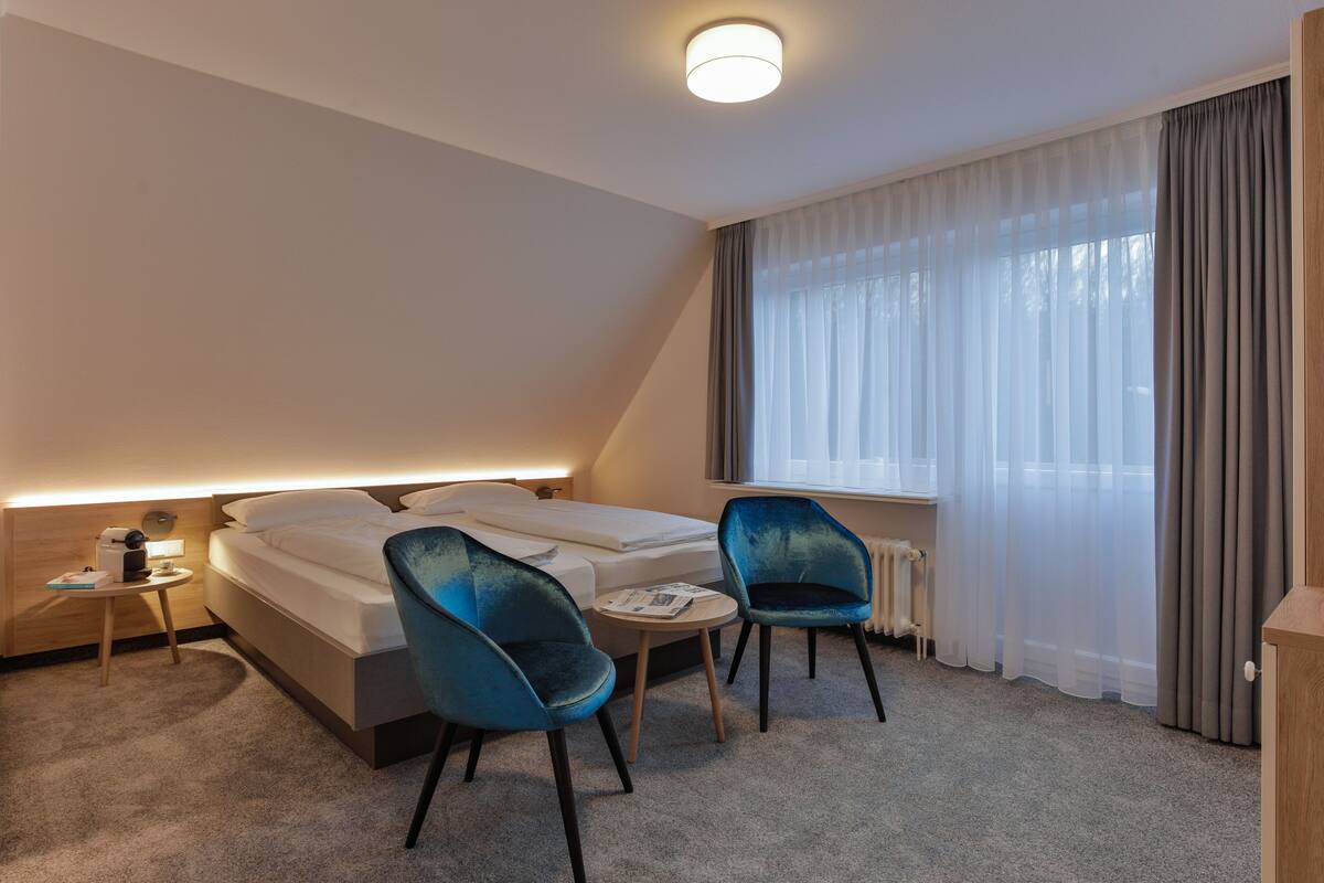 A hotel room with a bed, two chairs, a small table, a nightstand, and a window with curtains.