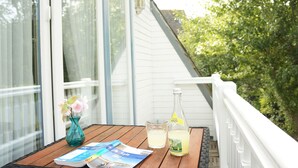 A wooden table with a magazine, a glass of water, and a bottle of lemonade on a balcony with white railings and a view of trees. - Hotel am Südwäldchen (Sylt)