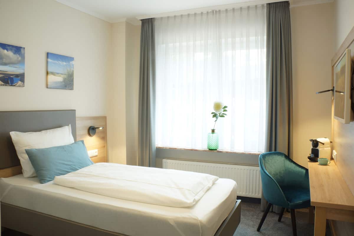 A hotel room with a bed, a desk, a chair, a window with curtains, and a radiator.