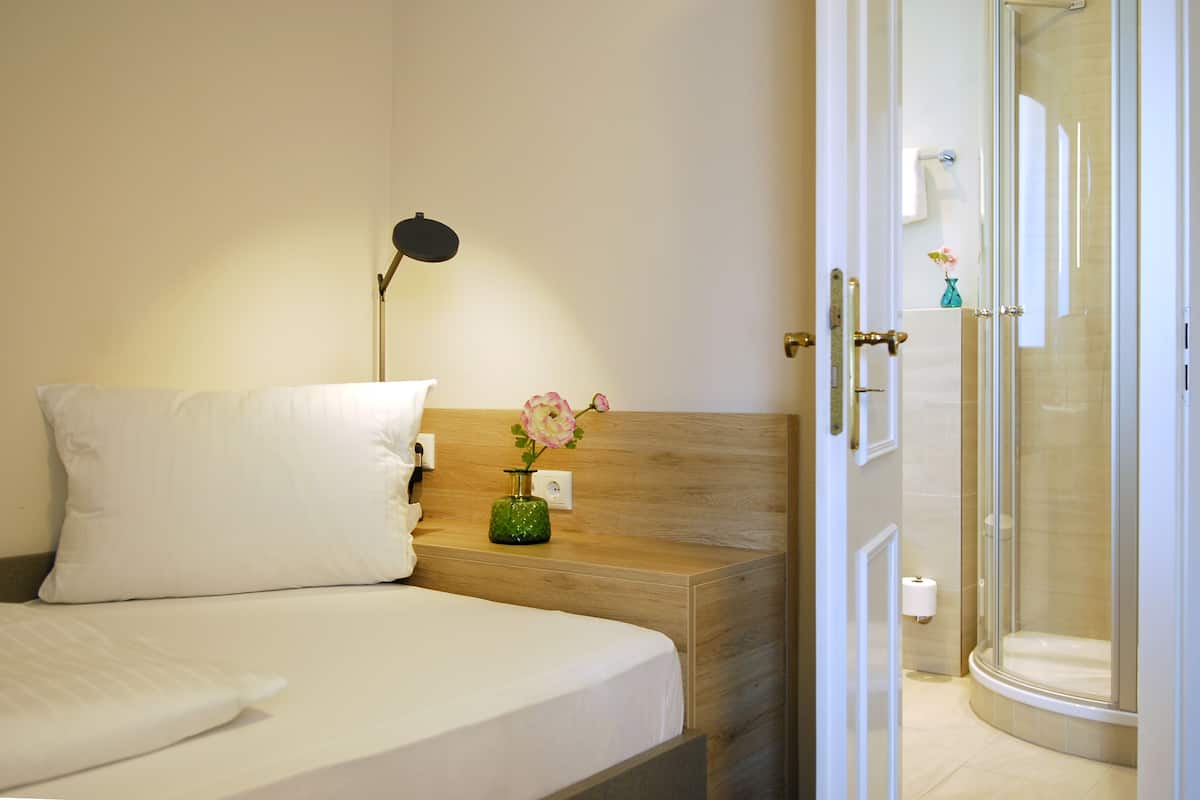 A hotel room with a bed, a bedside lamp, a vase with flowers, and a glass-enclosed shower area.