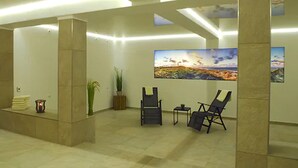 A modern lobby with a high ceiling, recessed lighting, and a large landscape painting on the wall. There are two lounge chairs and a small table. - Hotel am Südwäldchen (Sylt)