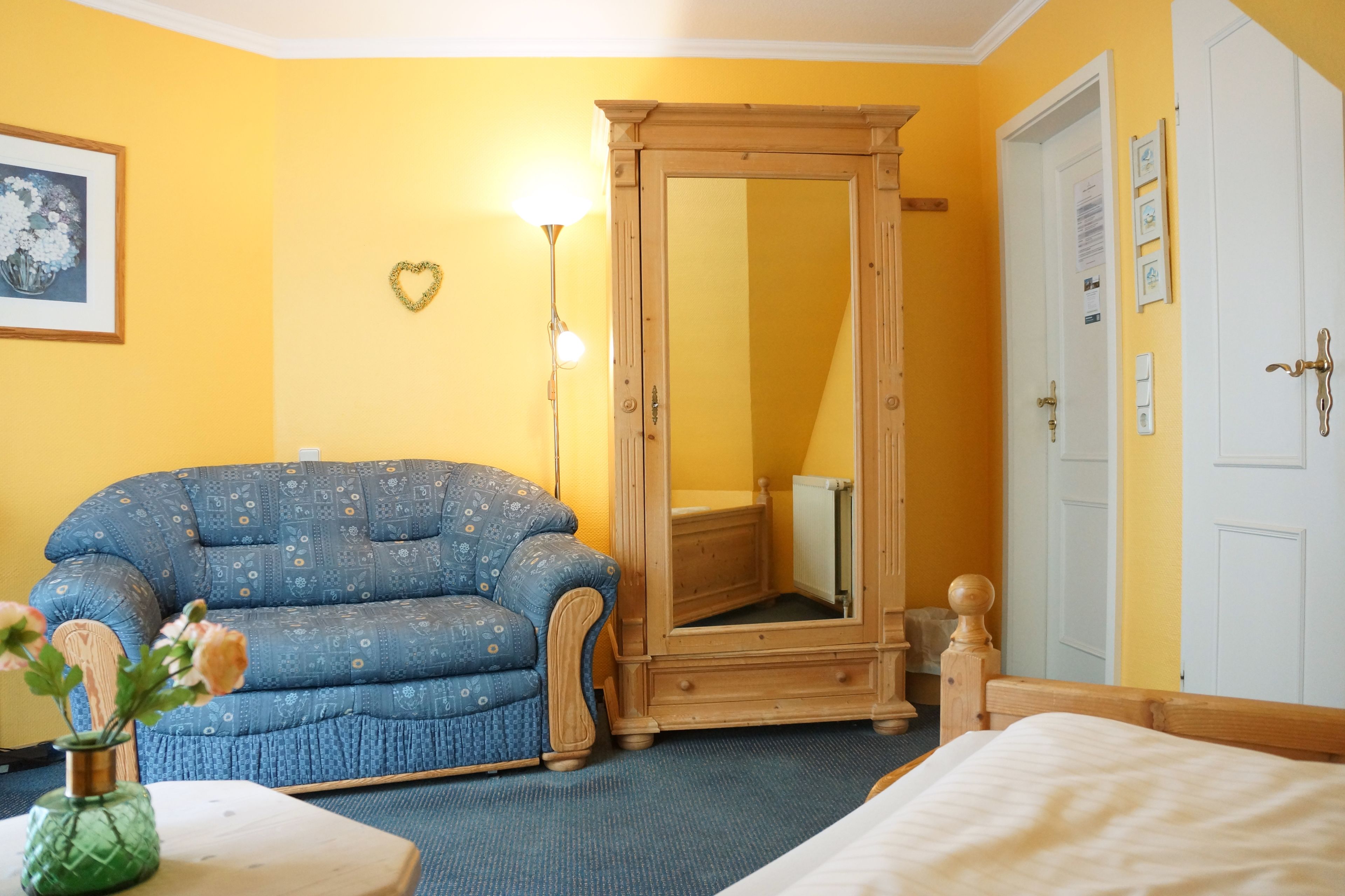 Een kamer met een blauwe sofa met patroon, een houten garderobe, een spiegel en een bed.