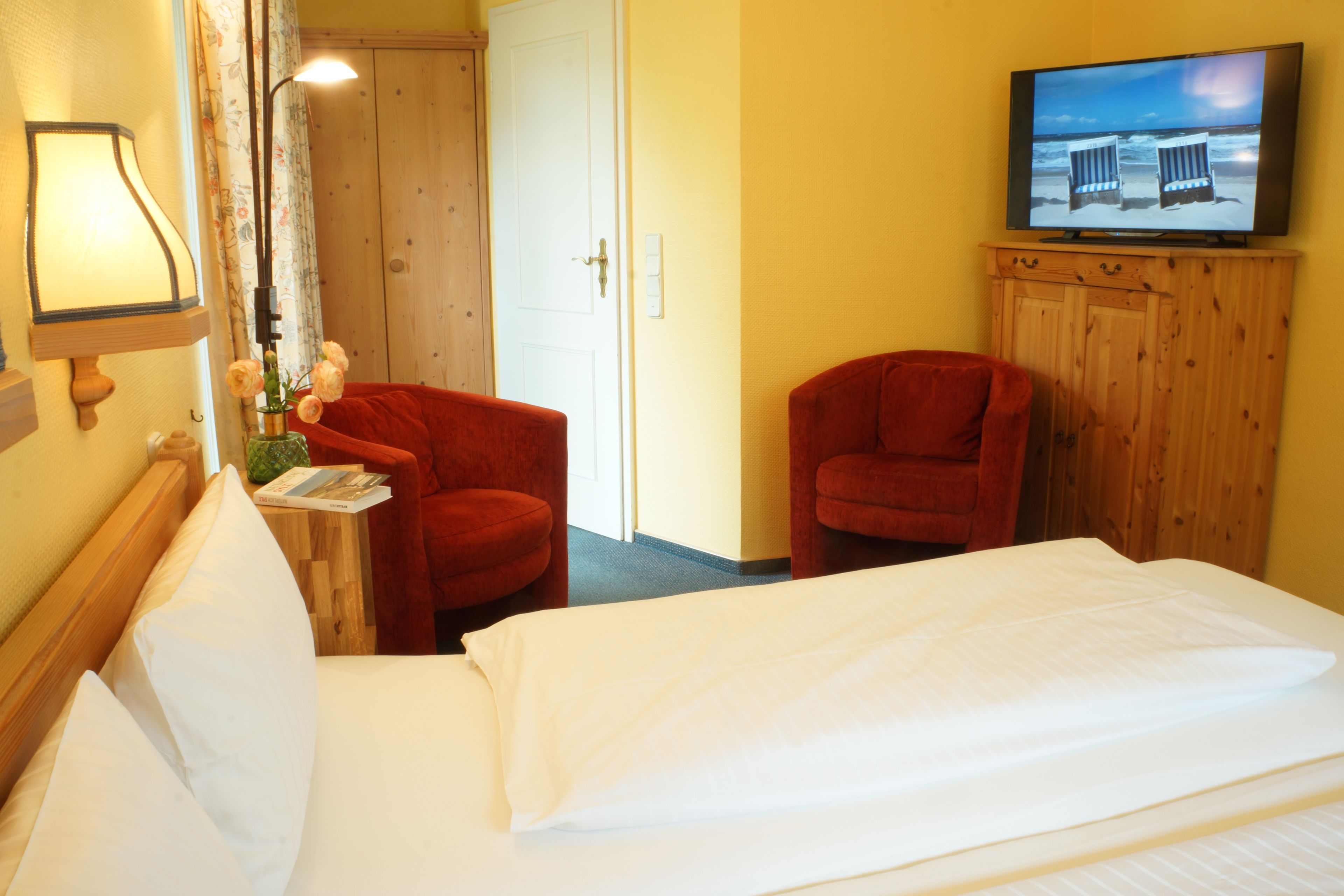 Une chambre d’hôtel avec un lit, deux fauteuils rouges, un téléviseur posé sur une commode en bois, une lampe et un vase avec des fleurs.