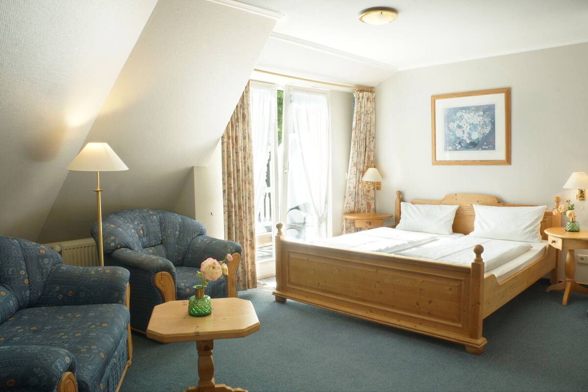 A bedroom with a wooden bed, two blue patterned armchairs, a small table with a vase, and a window with curtains.