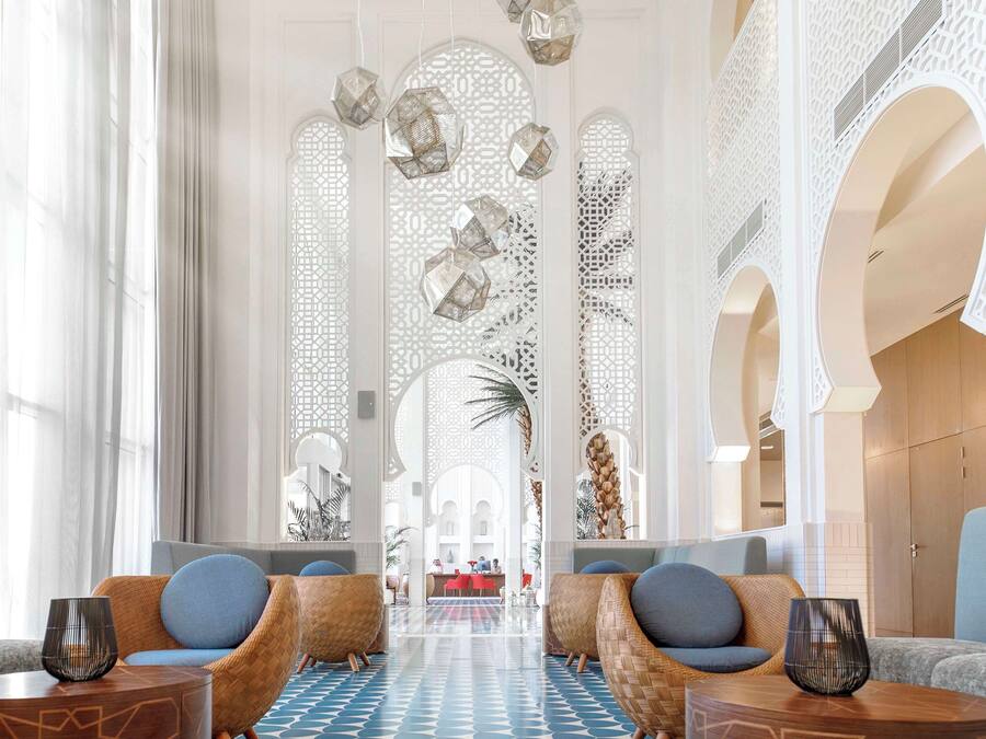 Um hall amplo com piso xadrez, áreas de estar com almofadas e luminárias pendentes decorativas.