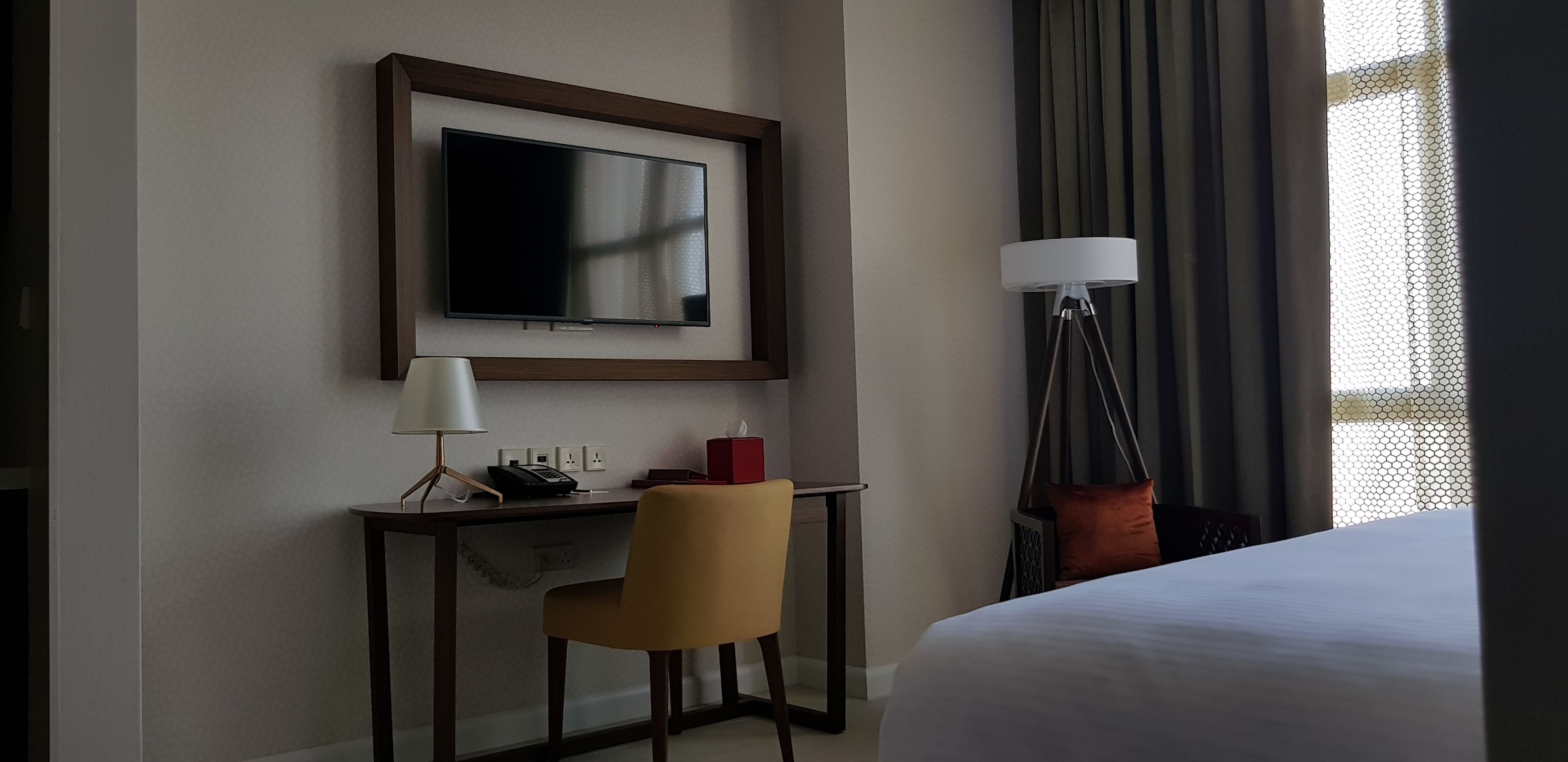 A hotel room with a bed, a desk with a lamp, a chair, a television mounted on the wall, and a floor lamp.