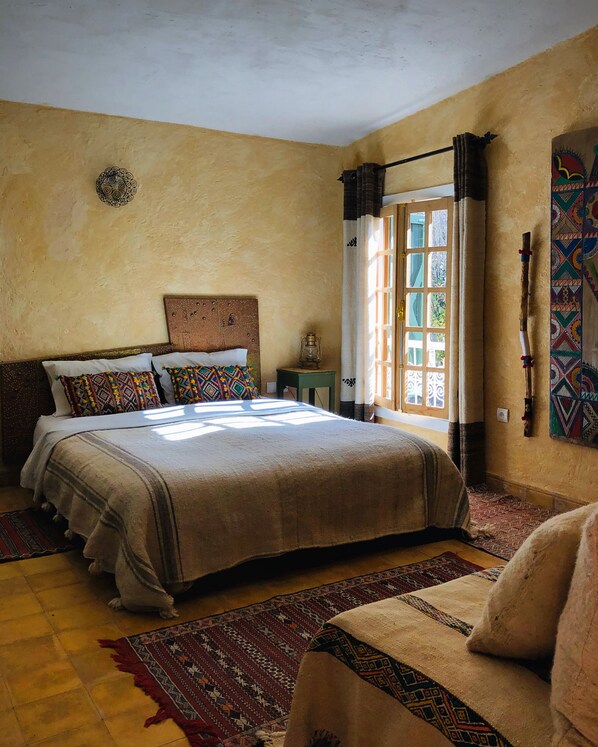 A bedroom with a large bed, a nightstand, a window with curtains, and a wall-mounted decoration. - Gite Dayet Aoua (Dayat Aoua)