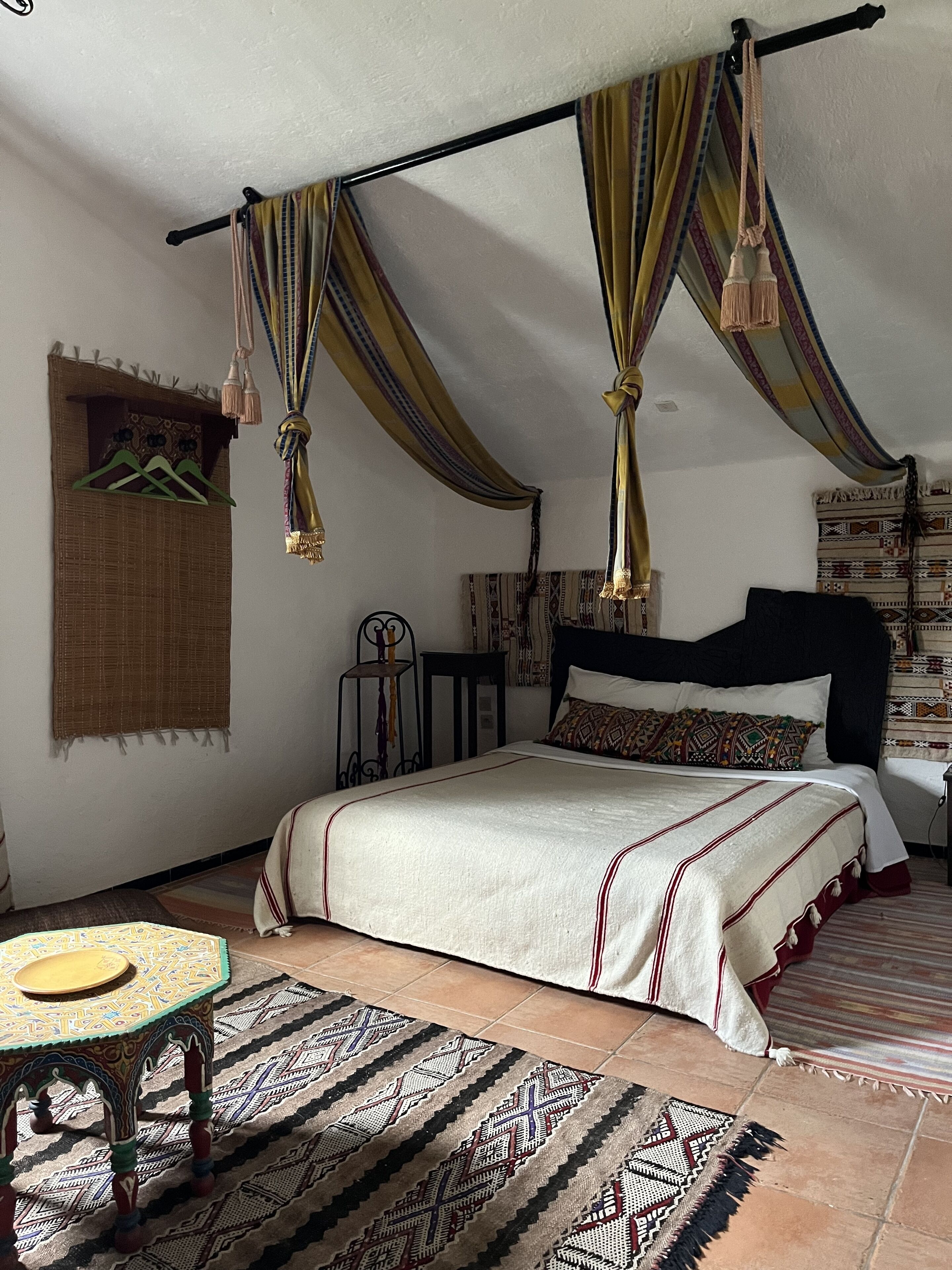A bedroom with a bed, a hanging chair, a small table, and patterned rugs.
