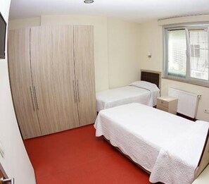 Un dormitorio pequeño y de una sola habitación con alfombra roja, dos camas, un armario y una ventana con persianas.