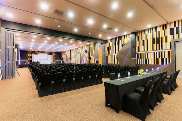 Un salón de conferencias con filas de sillas, una larga mesa y una pantalla de proyector.