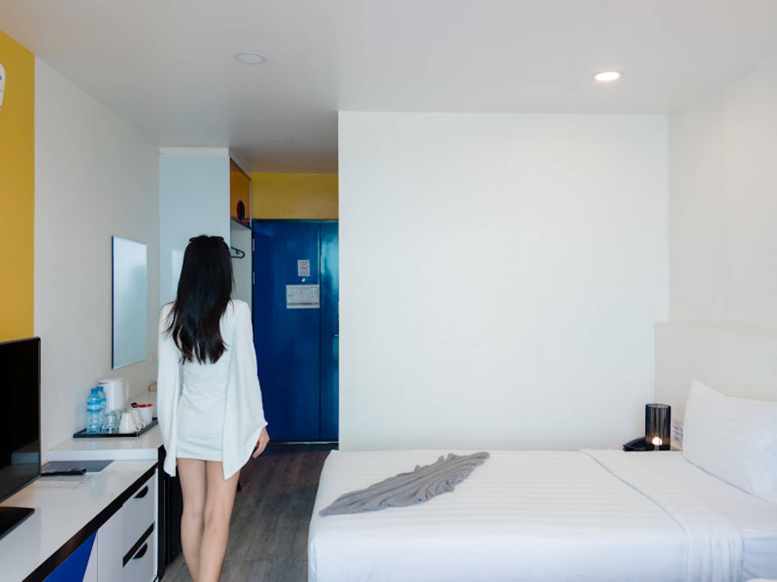 A hotel room with a bed, a desk with a chair, a mirror, and a blue door.