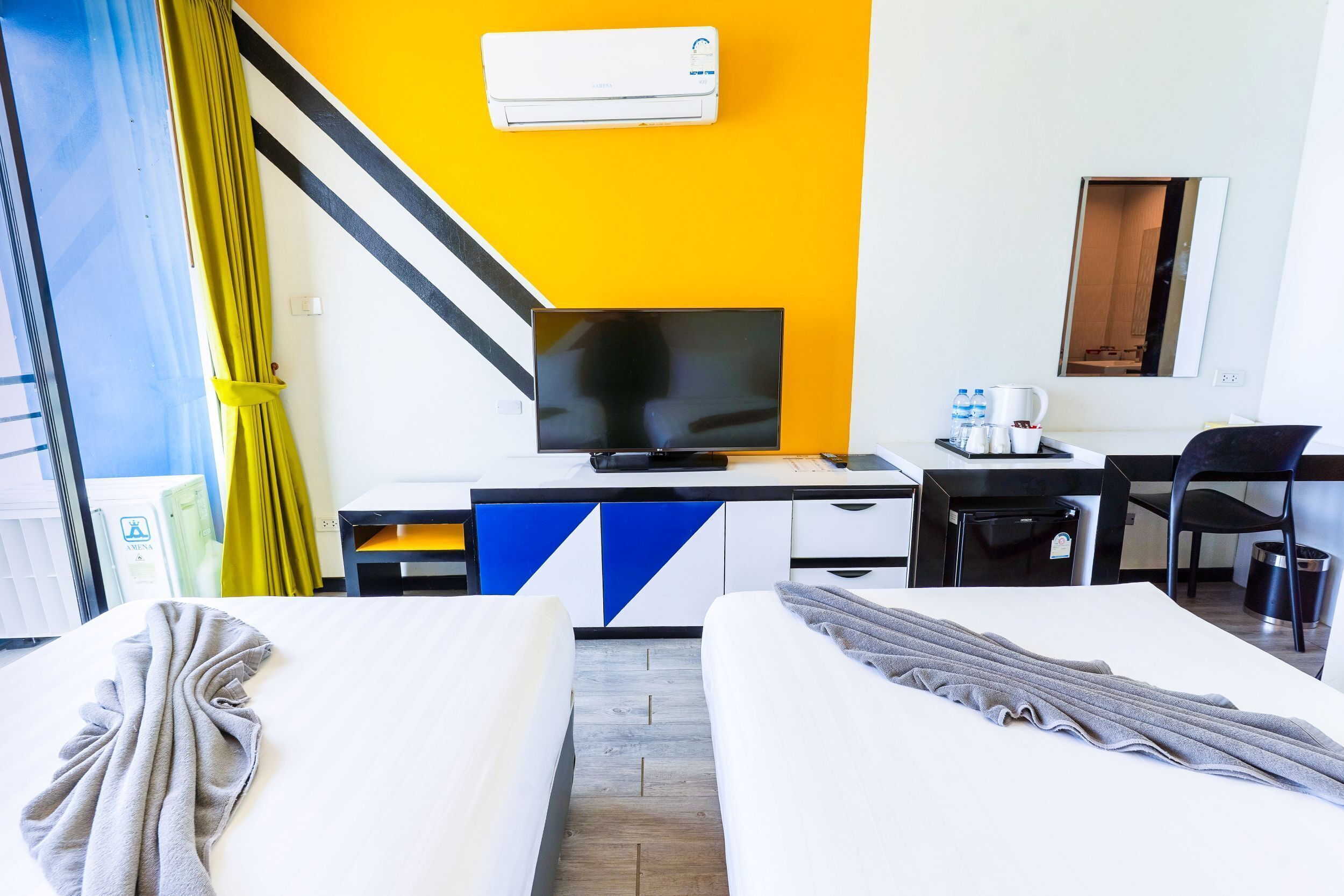 A hotel room with a bed, a desk with a television, a chair, and a small table with a kettle and cups.