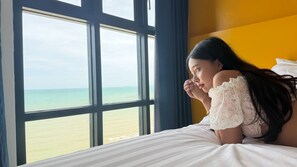 A room with a large window overlooking a beach, a bed with white bedding, and a yellow wall. - Play Phala Beach Rayong (Ban Chang)