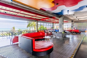 Un espace lounge moderne avec vue sur l’océan, comprenant des sièges aux couleurs rouge et noir, un sol à motifs et un plafond orné d’un décor coloré.