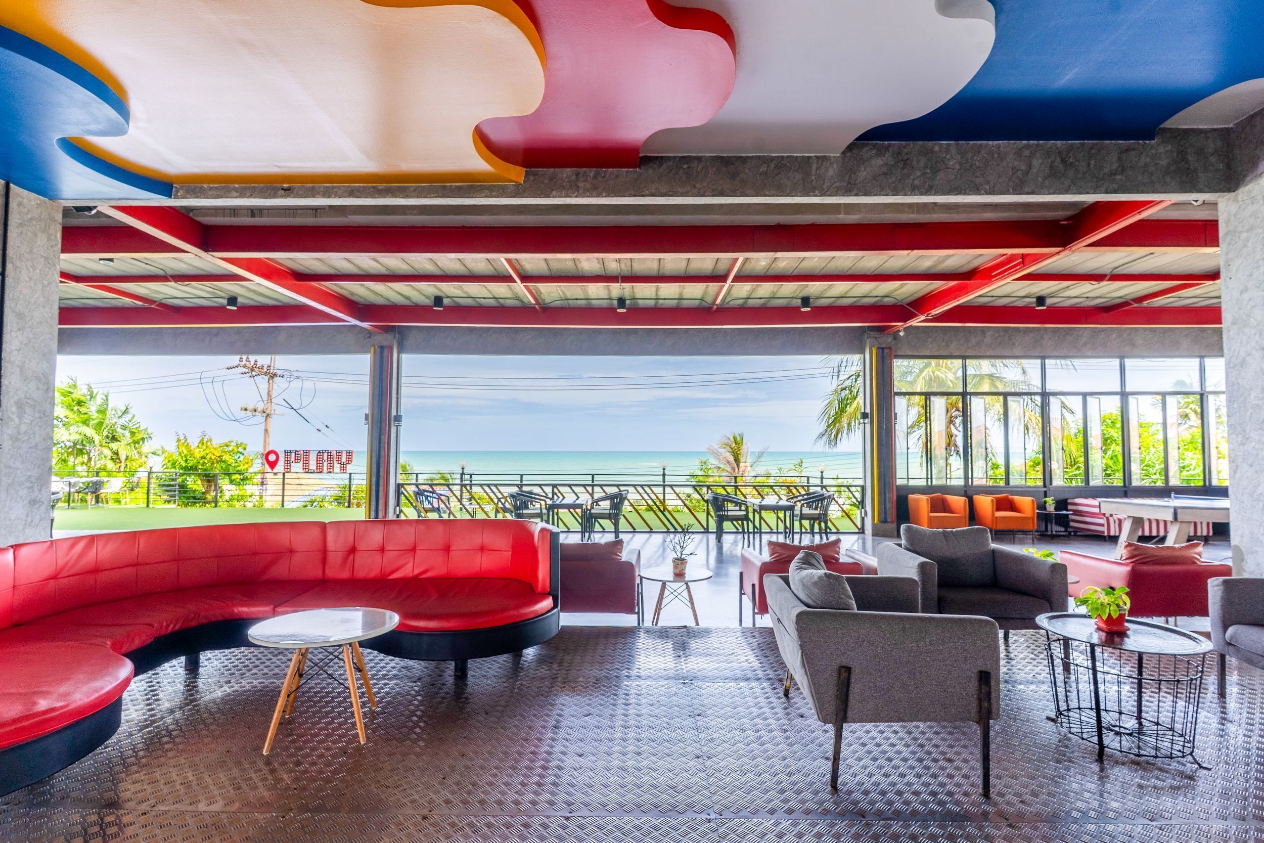 A modern lounge area with a curved red sofa, grey armchairs, and small tables. Large windows offer a view of the ocean.