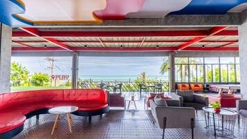 A modern lounge area with a curved red sofa, grey armchairs, and small tables. Large windows offer a view of the ocean.