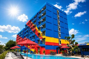 A multi-story hotel with a pool area, featuring a blue and red facade, multiple balconies, and red umbrellas. - Play Phala Beach Rayong (Ban Chang)