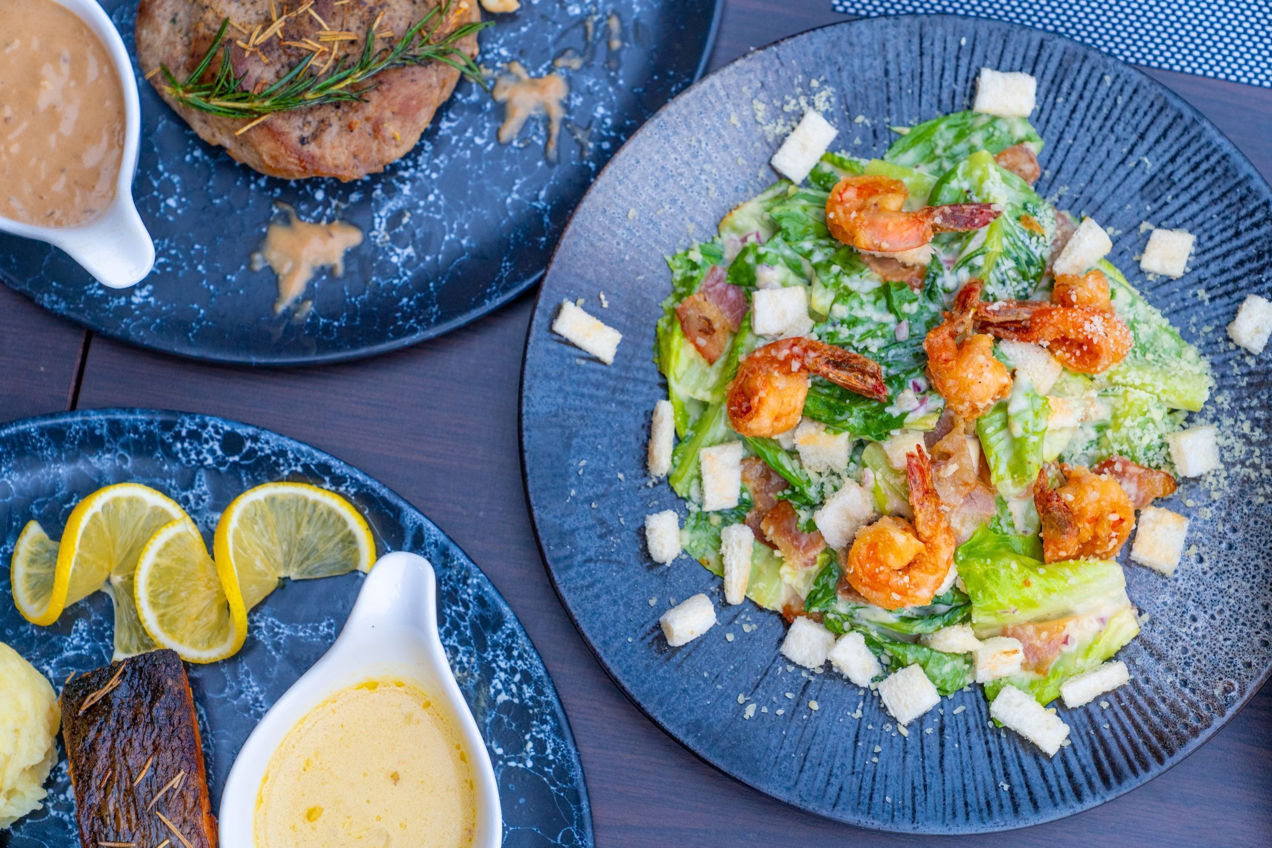Diversos pratos em travessas, incluindo uma salada com camarão e croutons, um pedaço de carne com alecrim e uma tigela de molho.
