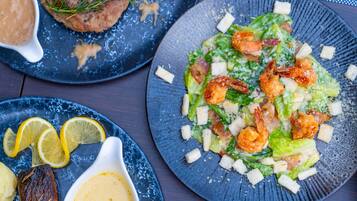 Diversos pratos em travessas, incluindo uma salada com camarão e croutons, um pedaço de carne com alecrim e uma tigela de molho.