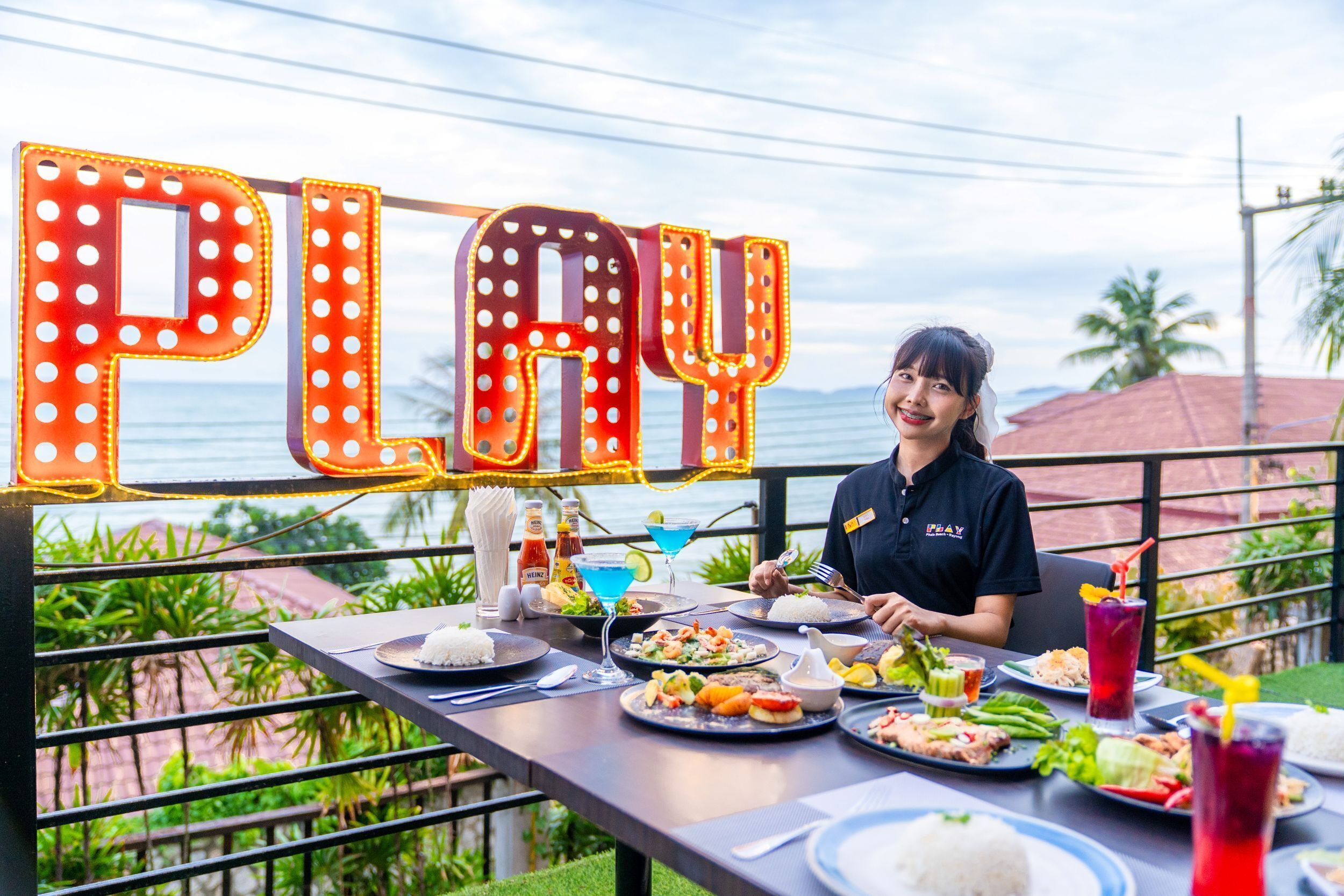 A person dining at an outdoor restaurant with a view of the ocean and a neon sign spelling 'PLAY' in the background.