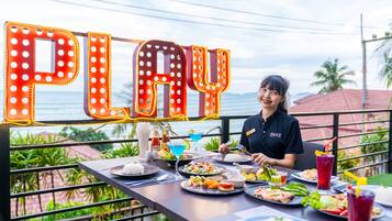 A person dining at an outdoor restaurant with a view of the ocean and a neon sign spelling 'PLAY' in the background.
