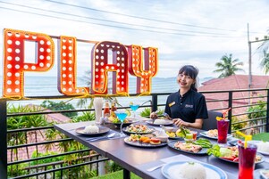 Una persona cenando en un restaurante al aire libre, con vista al océano y un letrero de neón que dice 'PLAY' al fondo.