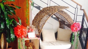 A cozy corner with a wicker sofa, a small wooden shelf with decorative items, and a large potted plant. - Lindi Hotel Baguio (Baguio)
