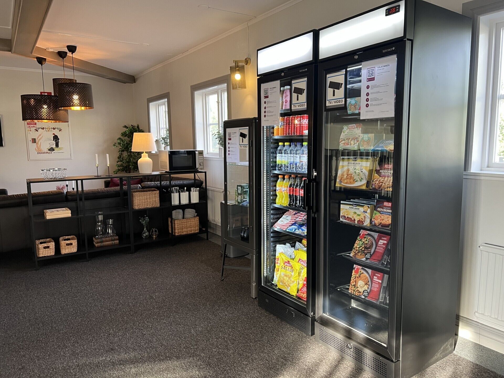 Eine Hotelhalle mit einer Selbstbedienungsgetränkestation, einer Sitzecke inklusive Sofa und Tisch sowie einem mit Snacks gefüllten Kühlschrank.