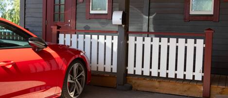 Un auto rojo estacionado frente a una casa con valla de piquete blanco, buzón y dos ventanas con contraventanas.