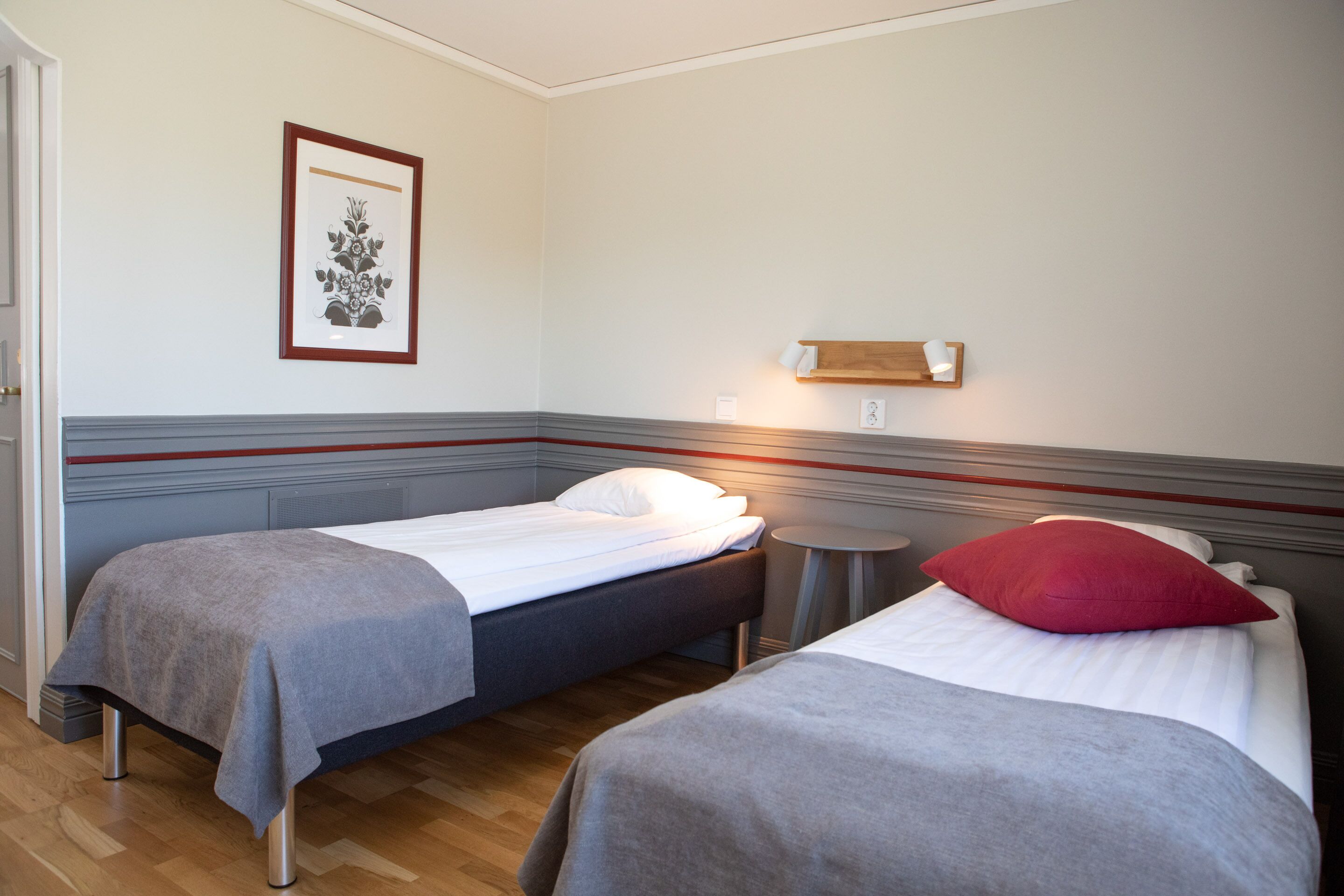 Habitación de hotel con dos camas individuales, una mesita redonda, una obra de arte en un marco en la pared y un estante de madera.